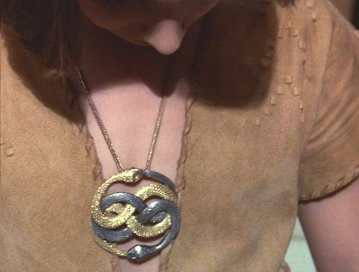 Medalhão dourado com duas serpentes entrelaçadas pendurado no peito de um personagem