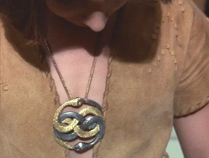 Medalhão dourado com duas serpentes entrelaçadas pendurado no peito de um personagem